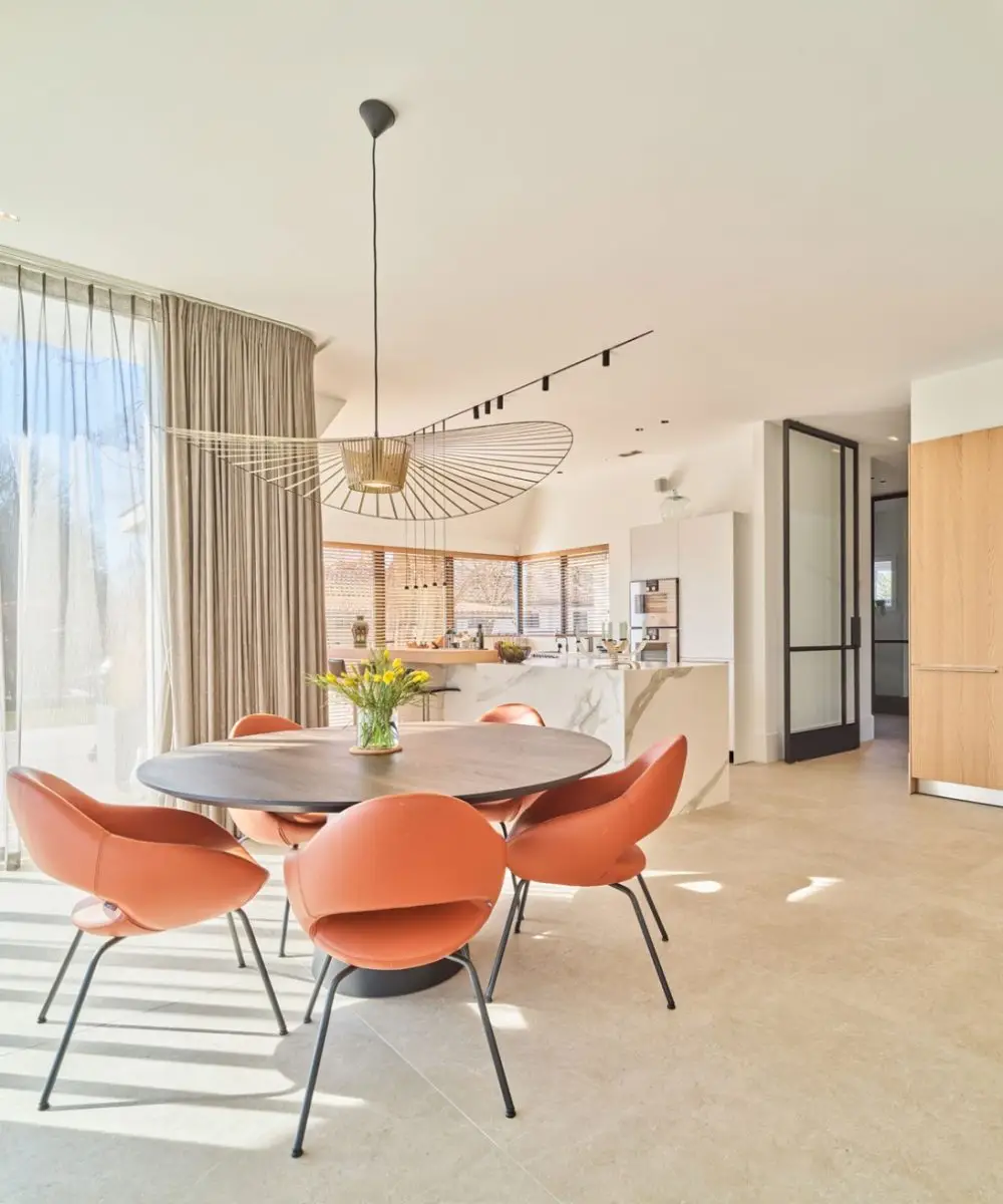 Lichte eetkeuken met ronde eettafel, oranje designstoelen, modern kookeiland en veel daglicht – Villa aan de Leede